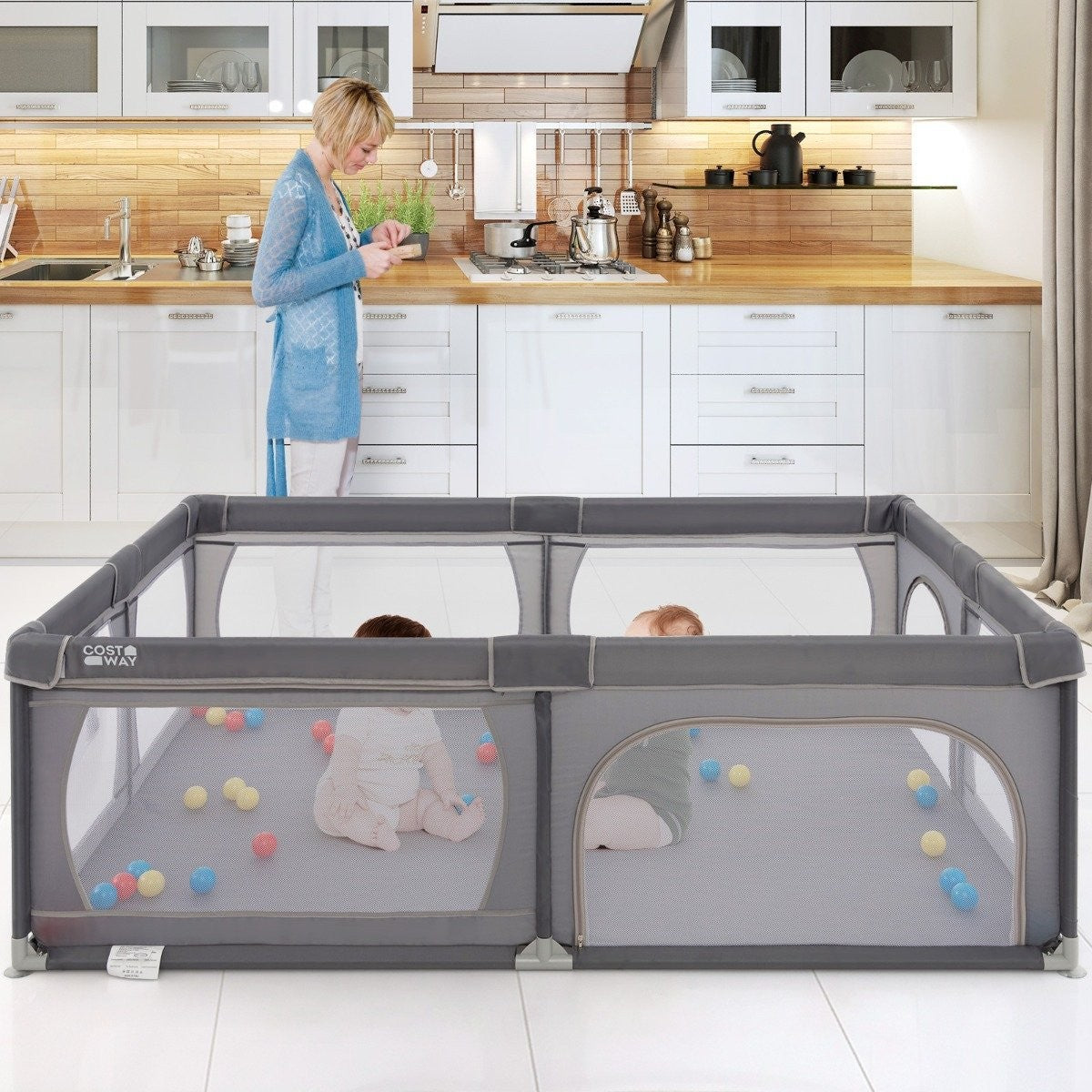 Una mujer en una cocina con un parque gris que contiene dos bebés y bolas coloridas. La cocina tiene gabinetes blancos.