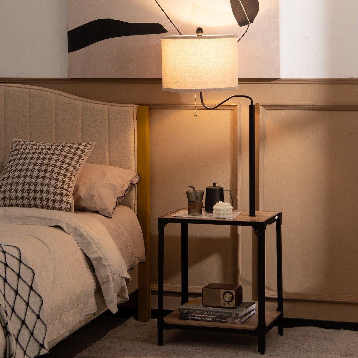 Escena de dormitorio con cama, lámpara de pie y mesita auxiliar. La cama tiene almohadas y una manta. La mesita tiene un radio y libros.