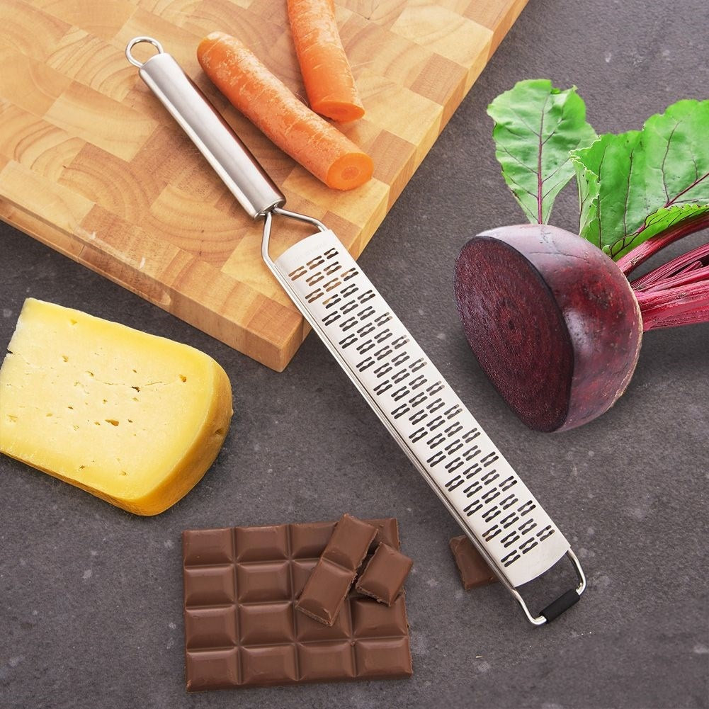 Un rallador, zanahorias, queso, chocolate y remolacha sobre una superficie oscura. También hay una tabla de cortar de madera.