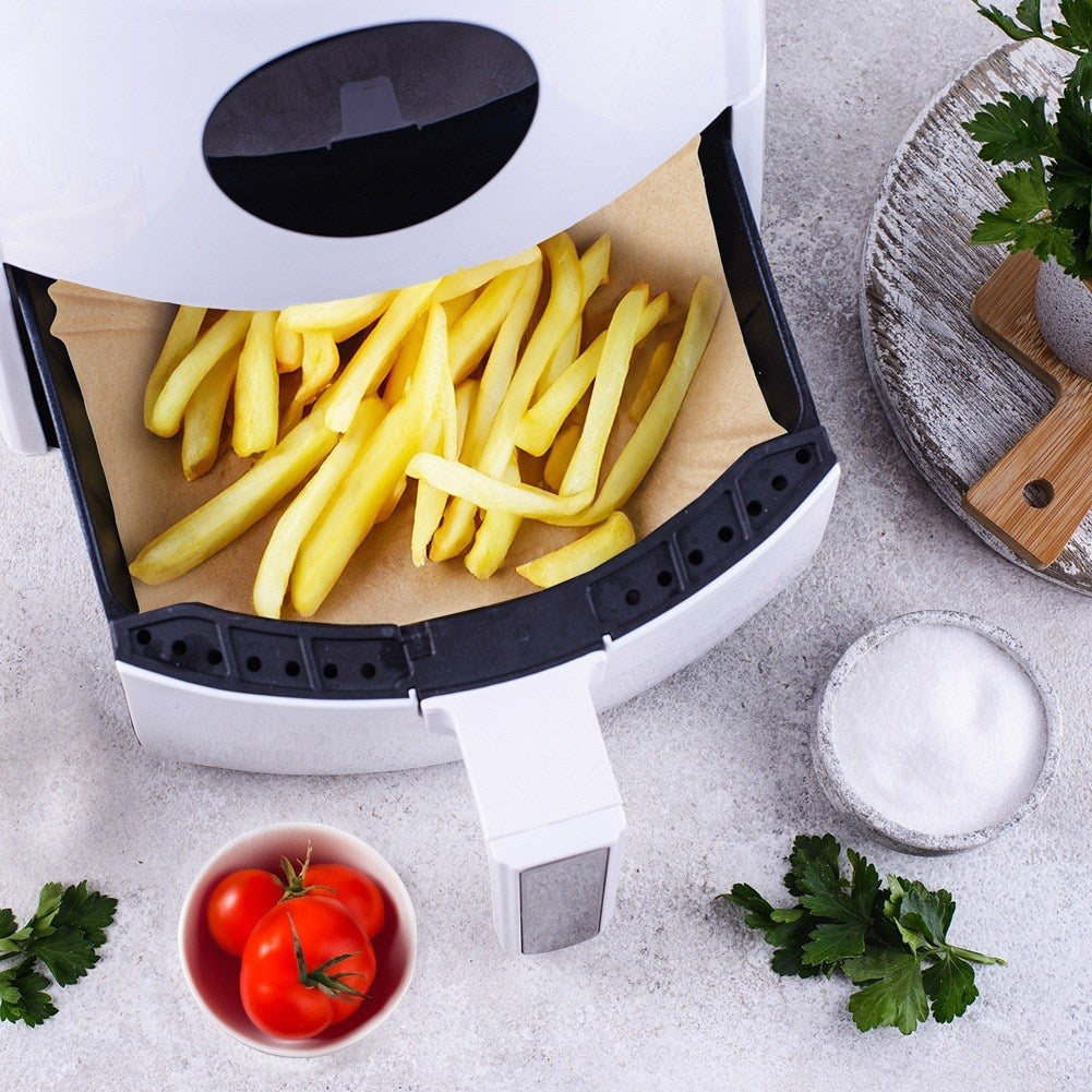 Una freidora de aire con papas fritas doradas, tomates, sal y perejil. Una tabla de madera con una planta al fondo.