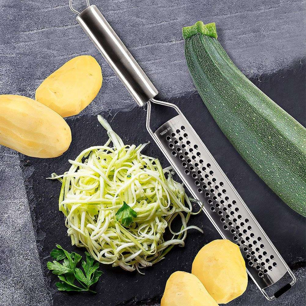 Un rallador, fideos de calabacín, calabacín, patatas y perejil sobre una pizarra negra. El rallador es de acero inoxidable.