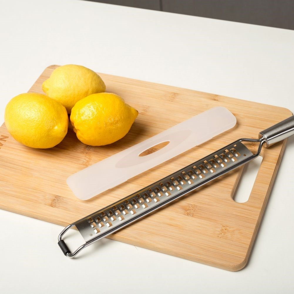 Tres limones amarillos, un rallador y una funda descansan sobre una tabla de cortar de madera. El fondo es blanco.