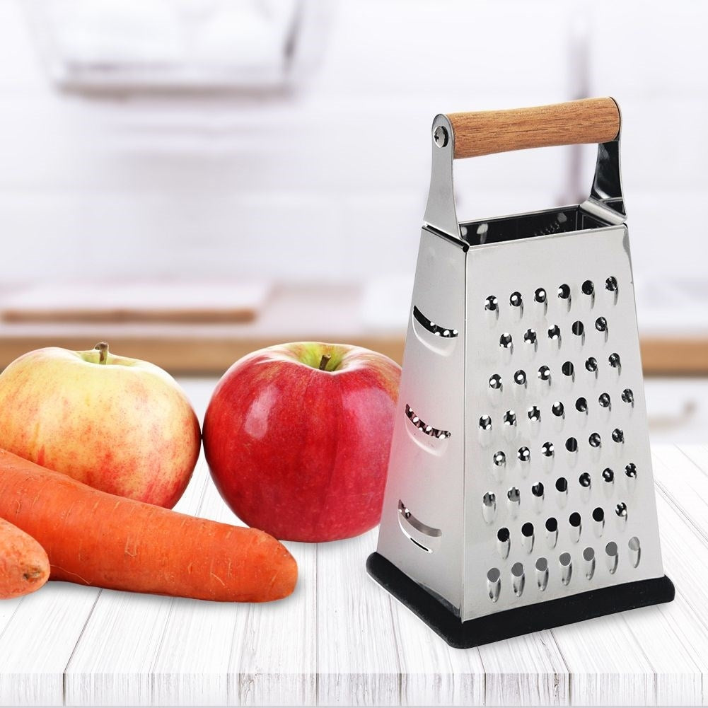Un rallador de metal con mango de madera junto a dos manzanas y zanahorias sobre una mesa de madera blanca. El fondo está borroso.