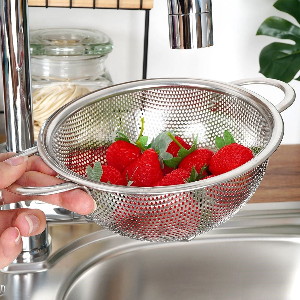 Una mano sostiene un colador lleno de fresas rojas. Encima hay un grifo y debajo un fregadero. Un tarro está en el fondo.