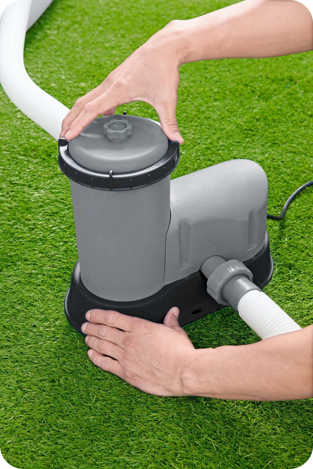 Manos montan un filtro de piscina gris. Mangueras blancas y una base negra son visibles sobre césped artificial verde.