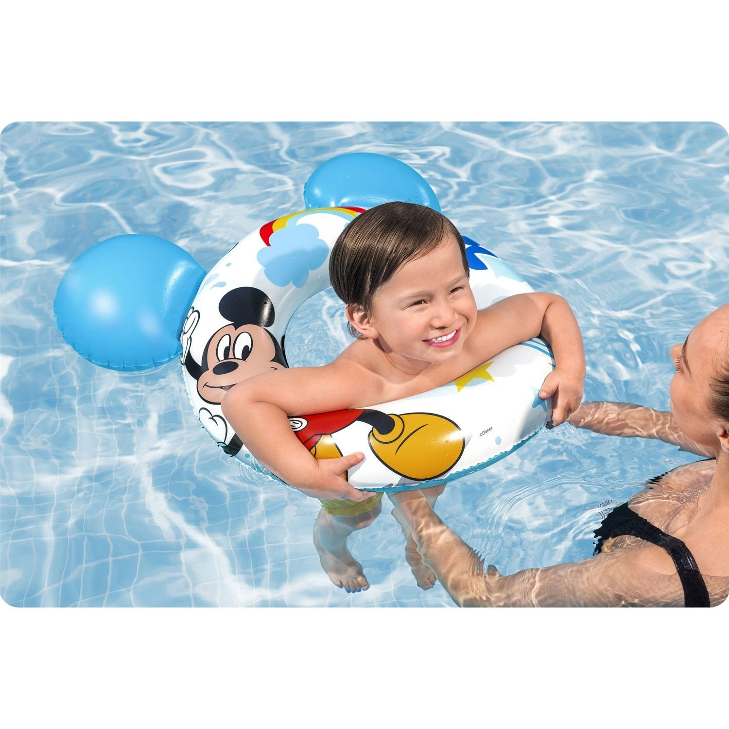 Un niño en un flotador de Mickey Mouse sonríe en una piscina. Un adulto sostiene al niño en el agua.