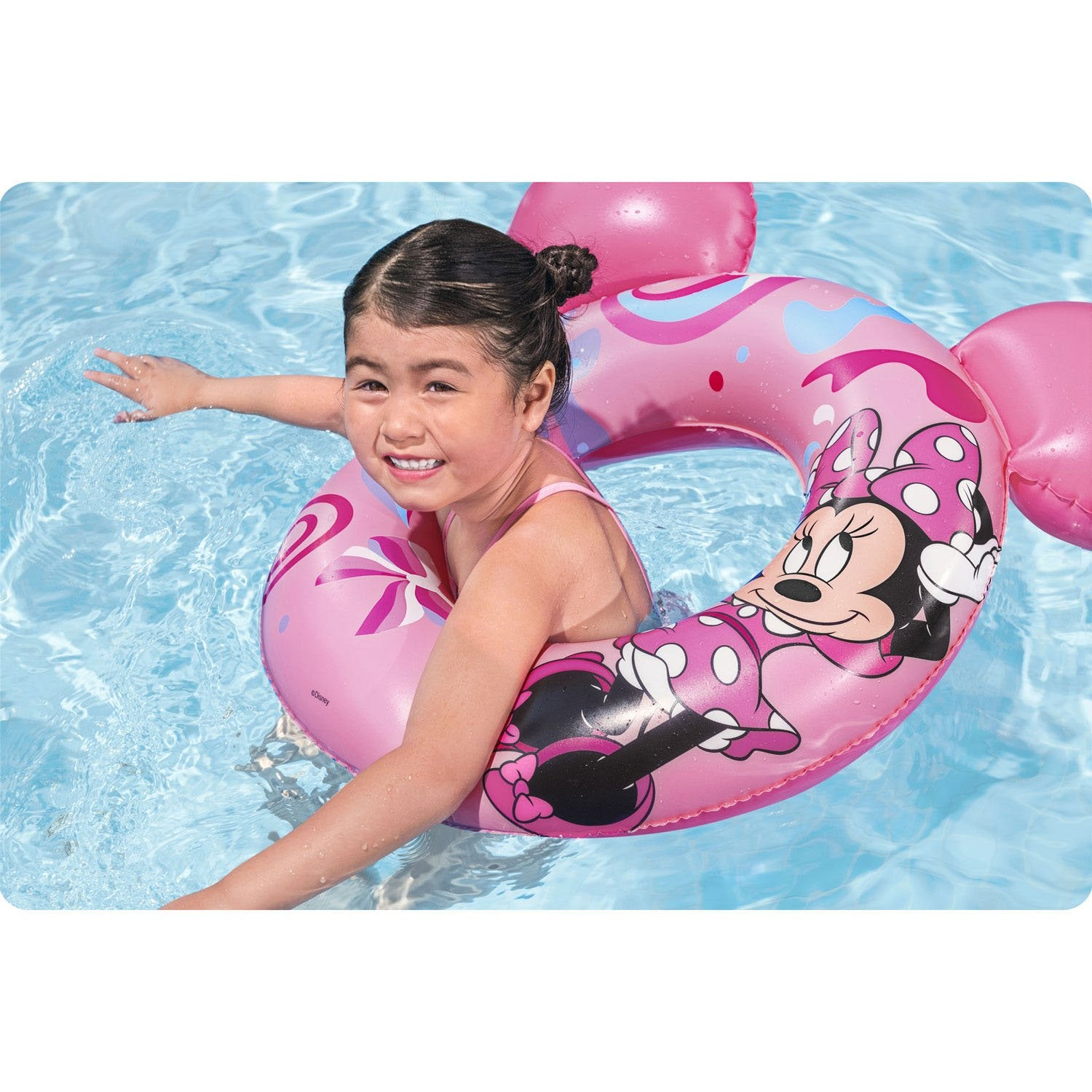 Una niña sonríe en una piscina, usando un flotador rosa de Minnie Mouse. El flotador tiene orejas de Mickey. El agua es azul.