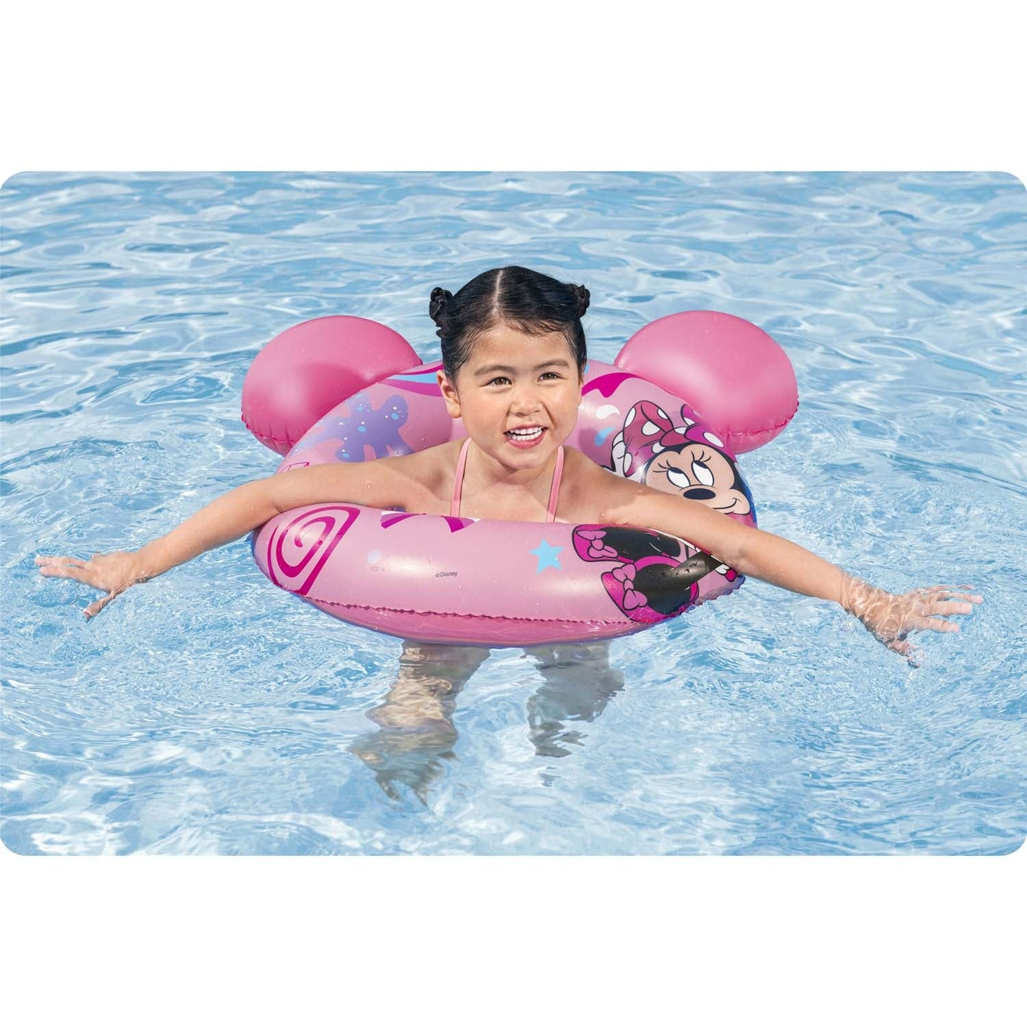 Una niña sonríe en una piscina, flotando en un flotador rosa de Minnie Mouse. El agua es azul. Lleva un traje de baño rosa.
