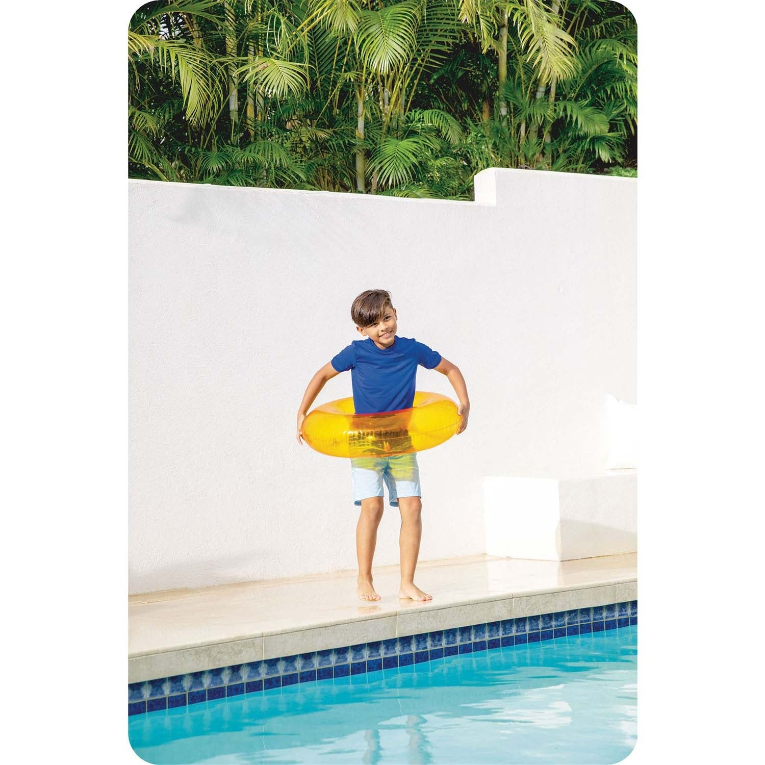 Un niño junto a la piscina, camisa azul y pantalones cortos, sosteniendo un flotador amarillo. Palmeras al fondo.