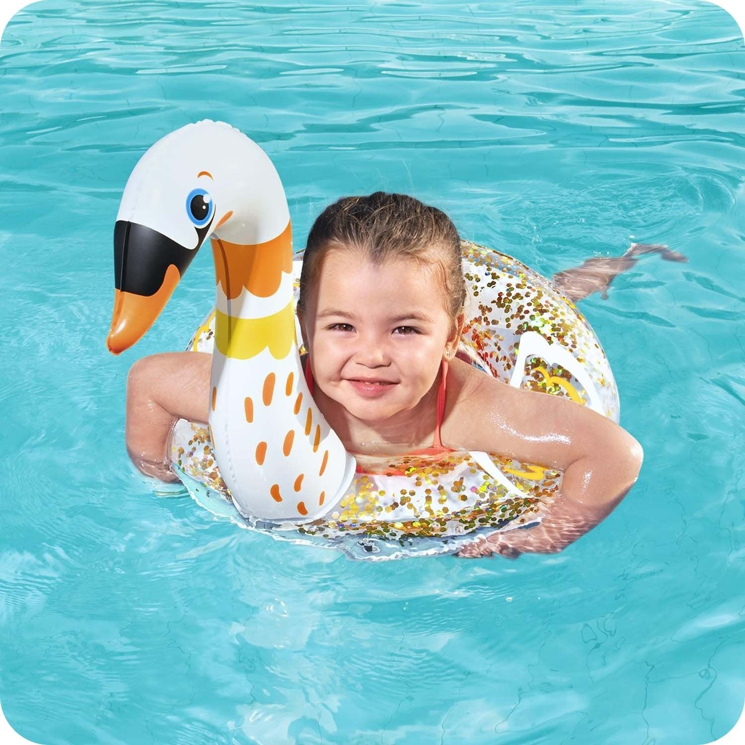 Una niña sonríe en una piscina mientras flota en un flotador inflable con forma de cisne. El agua es azul claro.