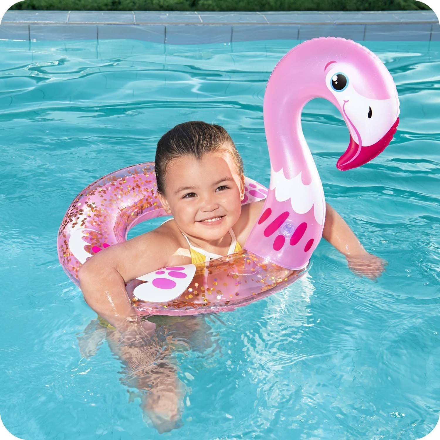 Una niña en una piscina sonríe, flotando en un flotador de flamenco rosa. El agua es azul. El flotador tiene purpurina dorada.