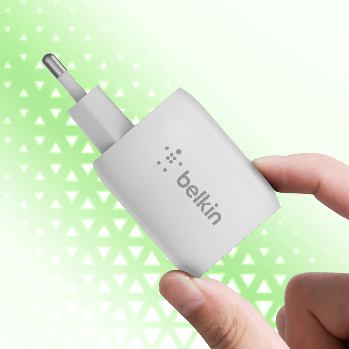 Un caricabatterie Belkin bianco tenuto dalle dita. Ha una spina e una porta USB. Lo sfondo è verde con triangoli bianchi.