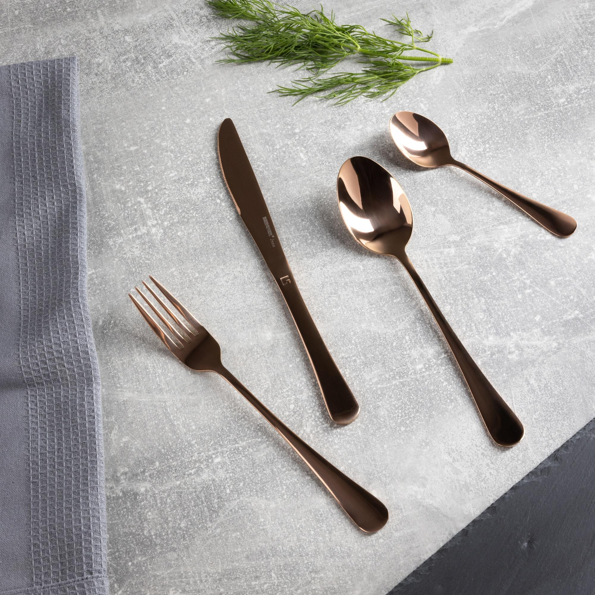 Juego de cubiertos de cobre sobre una superficie gris. Incluye tenedor, cuchillo, dos cucharas y ramita de eneldo, con tela gris.