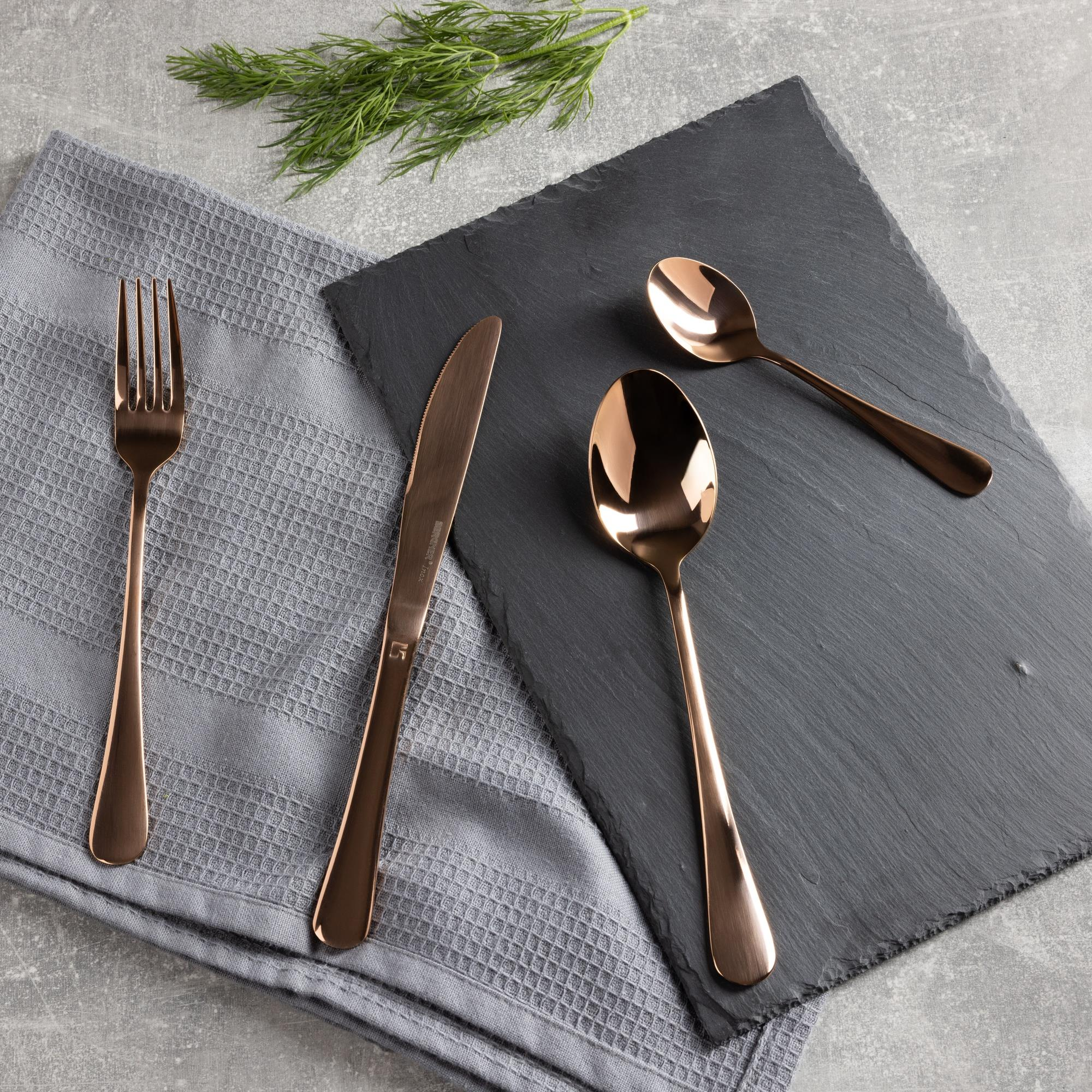 Cubertería de cobre sobre superficie gris: tenedor, cuchillo, cucharas y servilleta gris. Ramas de eneldo arriba a la izquierda.