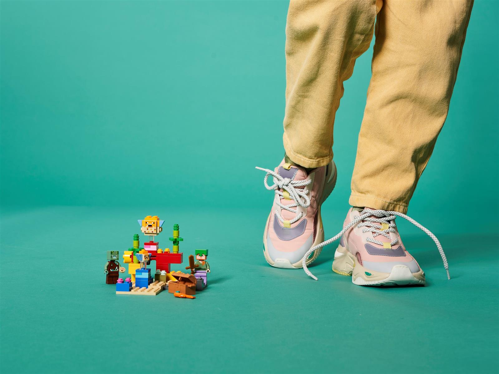 Eine Person mit gelber Hose und rosa Turnschuhen steht in der Nähe eines Lego-Sets auf türkisfarbenem Hintergrund.