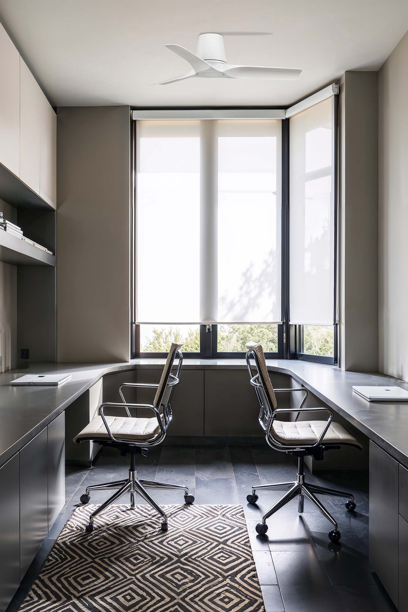 Büro mit zwei Stühlen, Schreibtisch und Fenster. Oben befindet sich ein weißer Deckenventilator. Ein Teppich mit geometrischem Muster liegt auf dem Boden.