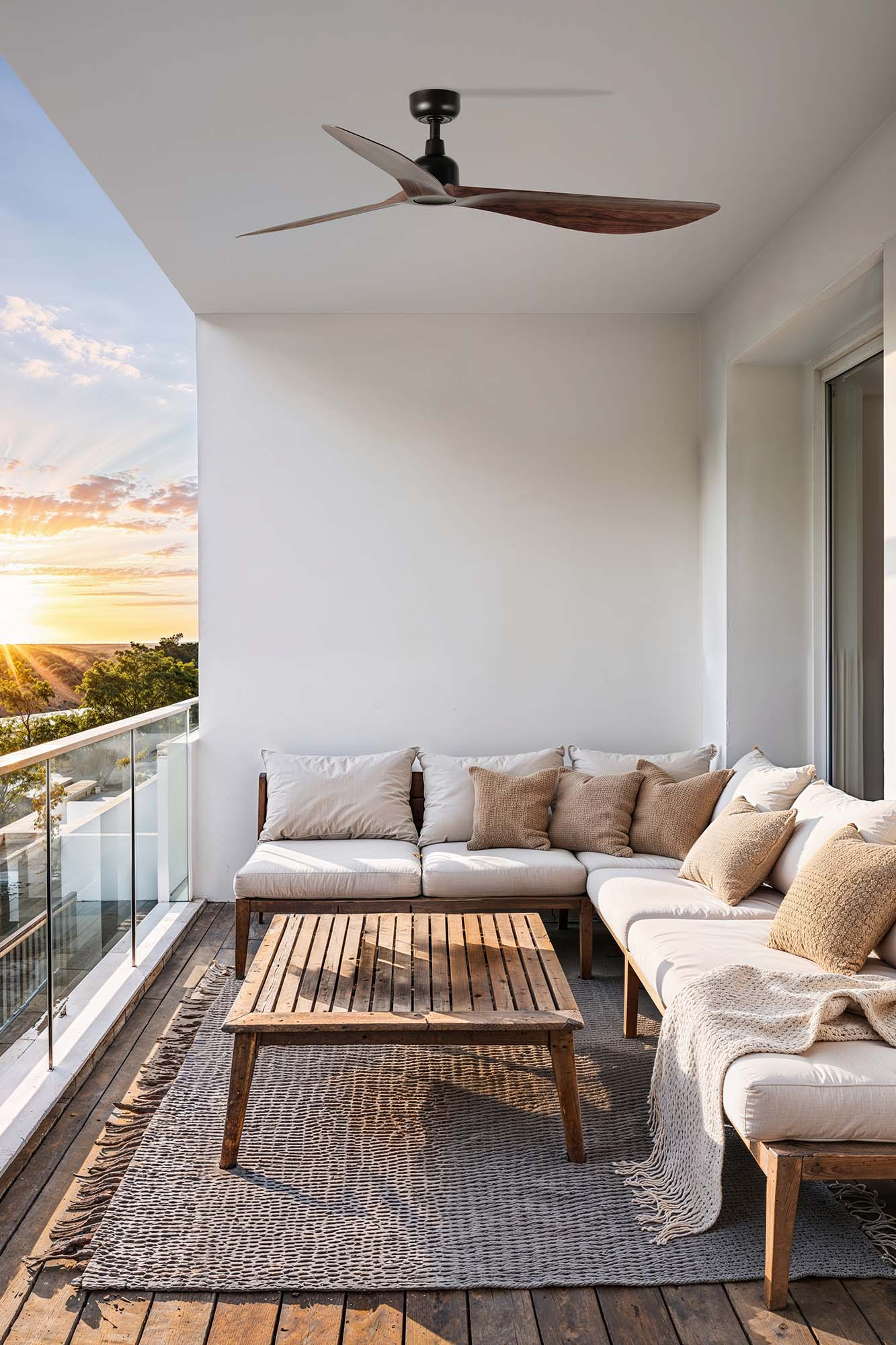 Balkon mit Deckenventilator, weißen Wänden, Holzmöbeln, Kissen, Teppich und Blick auf den Sonnenuntergang.