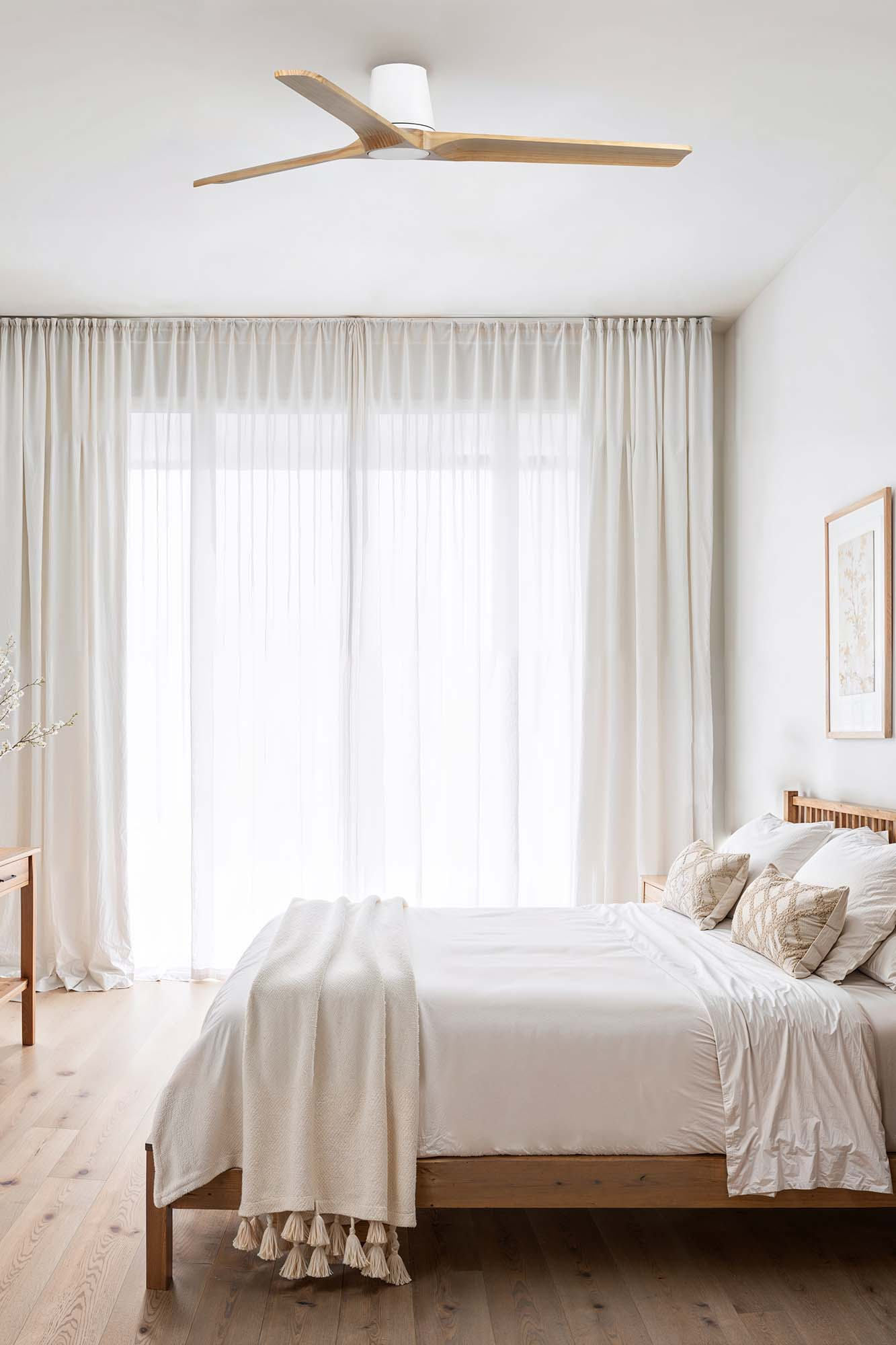 Schlafzimmer mit Bett, weißen Vorhängen, Holzboden und Deckenventilator. Das Bett hat Kissen und eine Decke. Weiches, natürliches Licht.