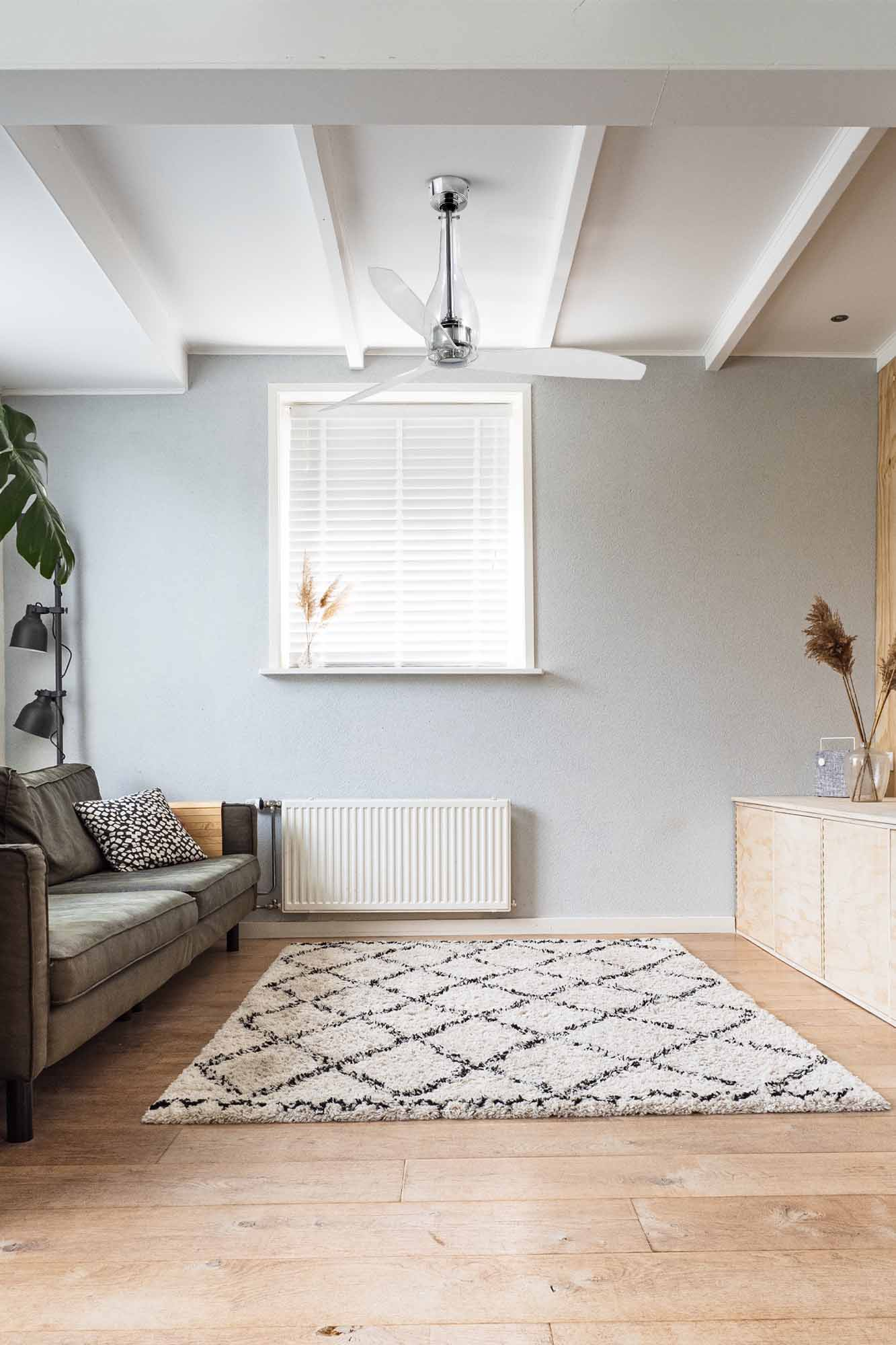 Wohnzimmer mit Sofa, Teppich, Deckenventilator und Fenster mit Jalousien. Heller Holzboden, graue Wände und weiße Decke.