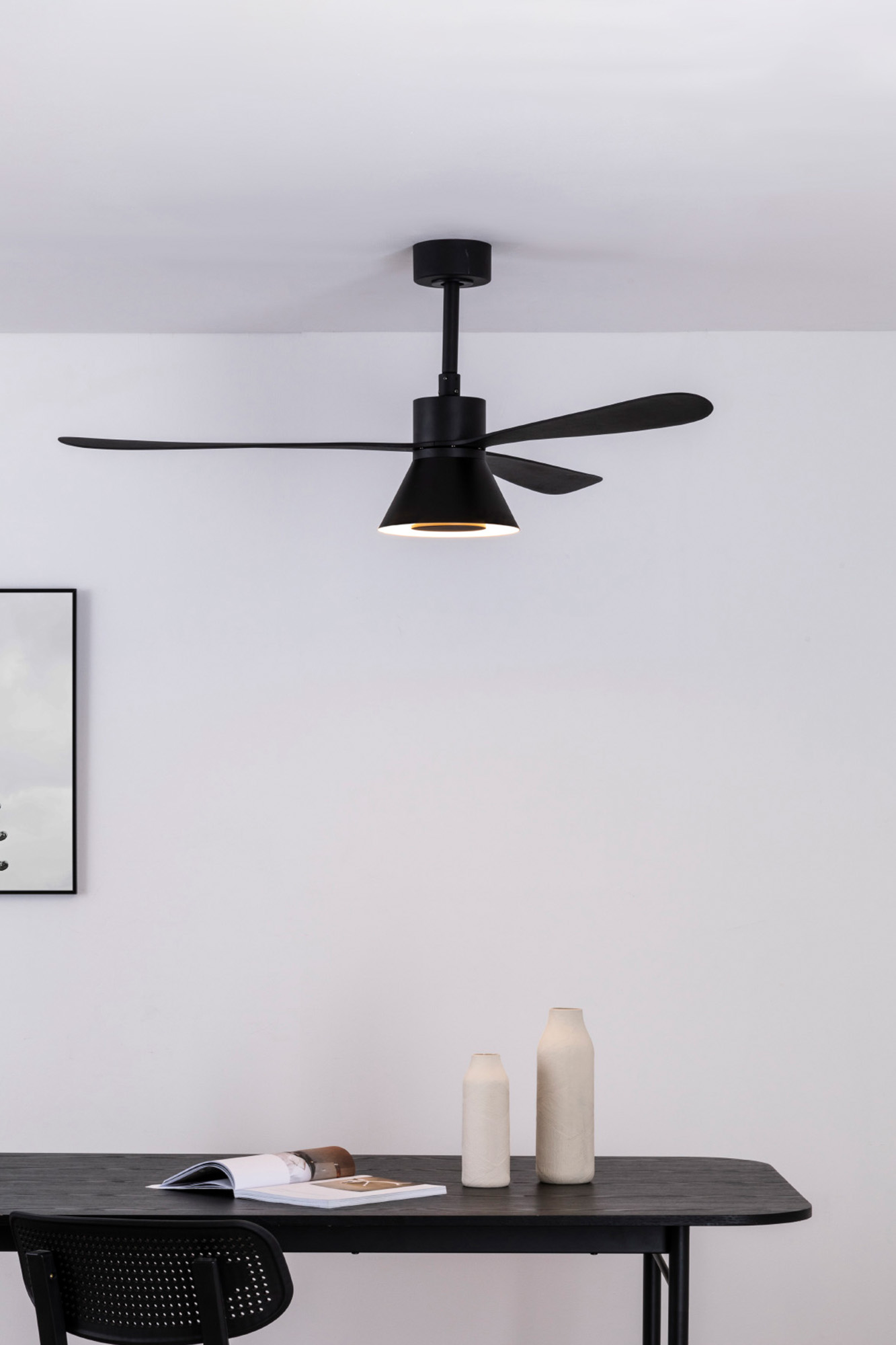 Schwarzer Deckenventilator über einem Tisch mit Vasen und einem Buch. Ein gerahmtes Bild hängt an der Wand.