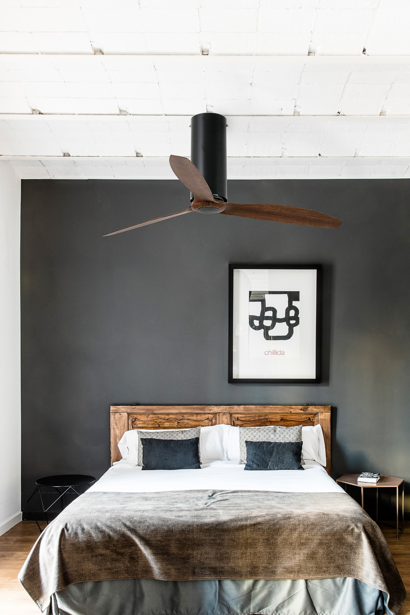 Schlafzimmer mit Bett, Kunst, Deckenventilator und dunkelgrauer Wand. Holz-Kopfteil und Beistelltisch. Weiße Decke und Boden.