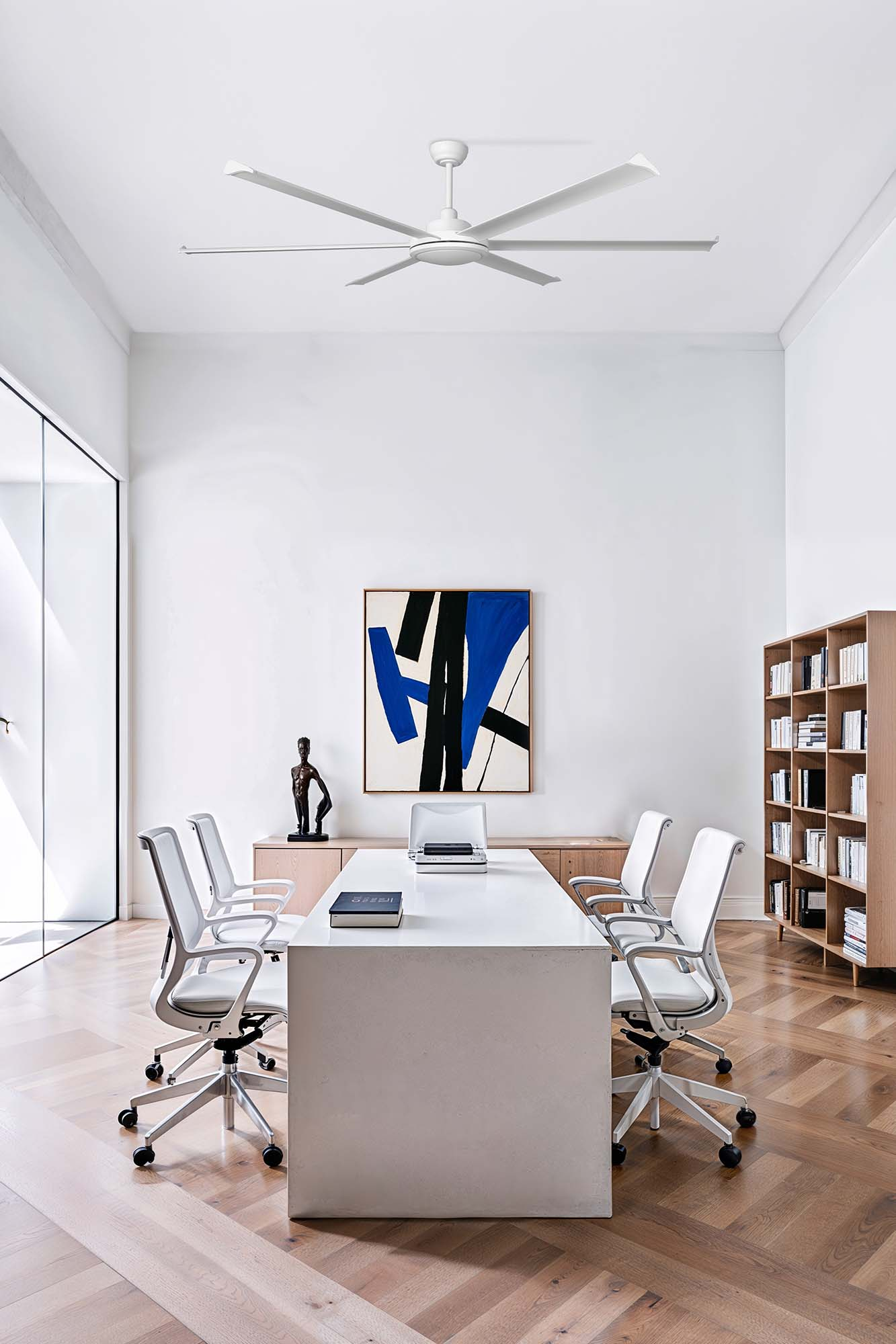 Ufficio moderno con pareti bianche, ventilatore a soffitto, grande tavolo, sedie, arte e pavimento in legno. Una statua è sul tavolo.