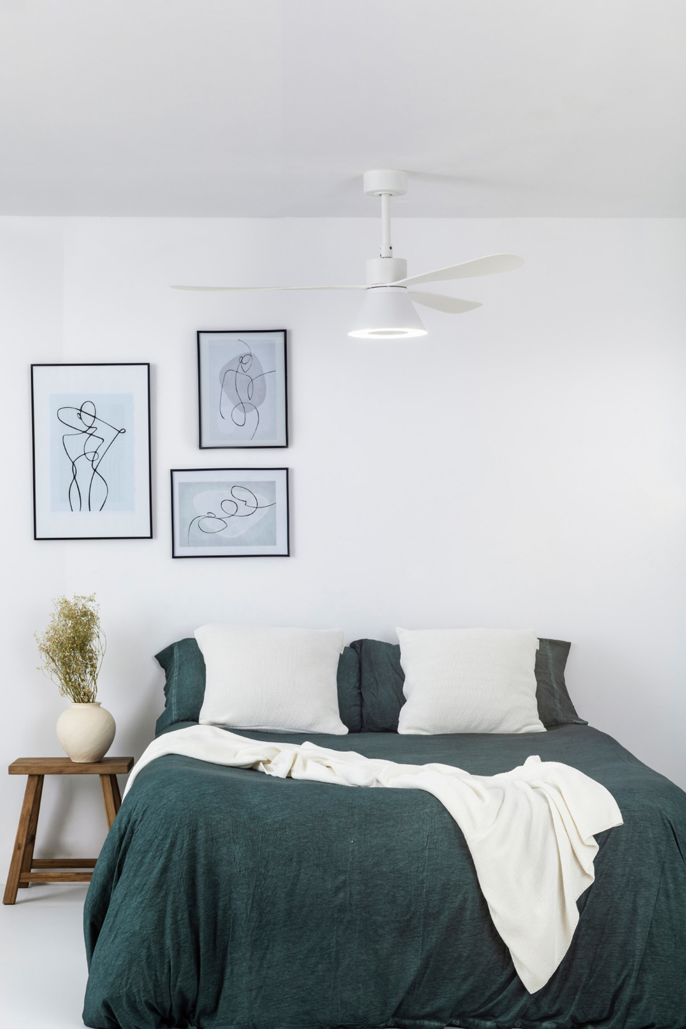 Schlafzimmer mit Bett, weißen Kissen, grüner Bettdecke, weißer Decke, Kunst und Deckenventilator. Eine Vase steht auf einem kleinen Holztisch.