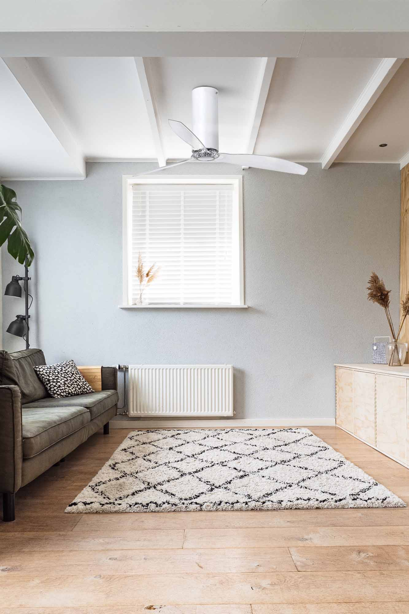Ein modernes Wohnzimmer mit Deckenventilator, Sofa, Teppich und Fenster mit Jalousien. Helle Holzböden und eine hellblaue Wand.