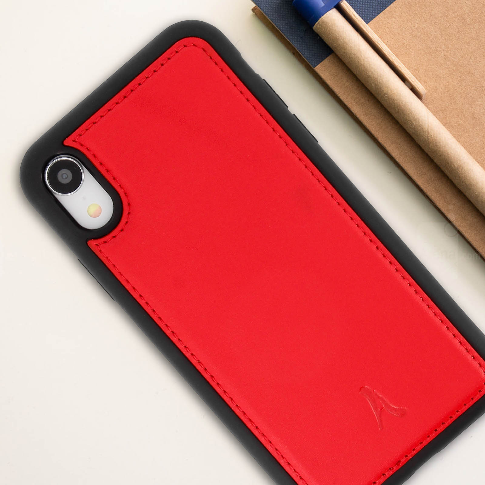 Custodia rossa per telefono su superficie bianca, con bordo nero, fotocamera e quaderno con penna sullo sfondo.