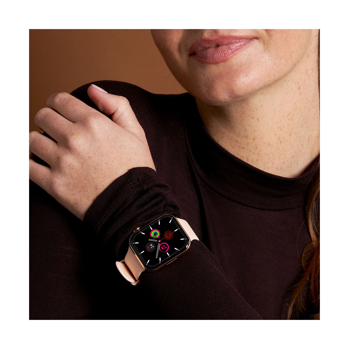 Une femme porte une montre connectée avec un bracelet rose. Le cadran affiche diverses icônes. Elle porte une chemise marron foncé à manches longues.