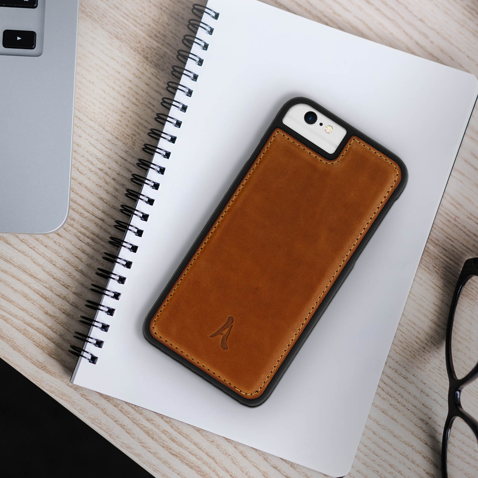 Una custodia per telefono in pelle marrone poggia su un taccuino bianco. Visibili anche un laptop e occhiali.