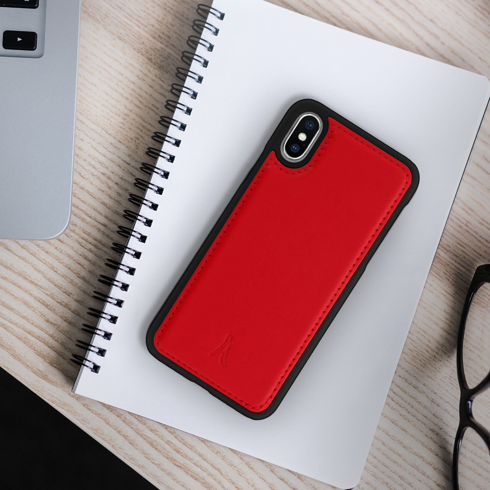 Custodia rossa per telefono su un taccuino. Un laptop e occhiali sono parzialmente visibili su un tavolo di legno.