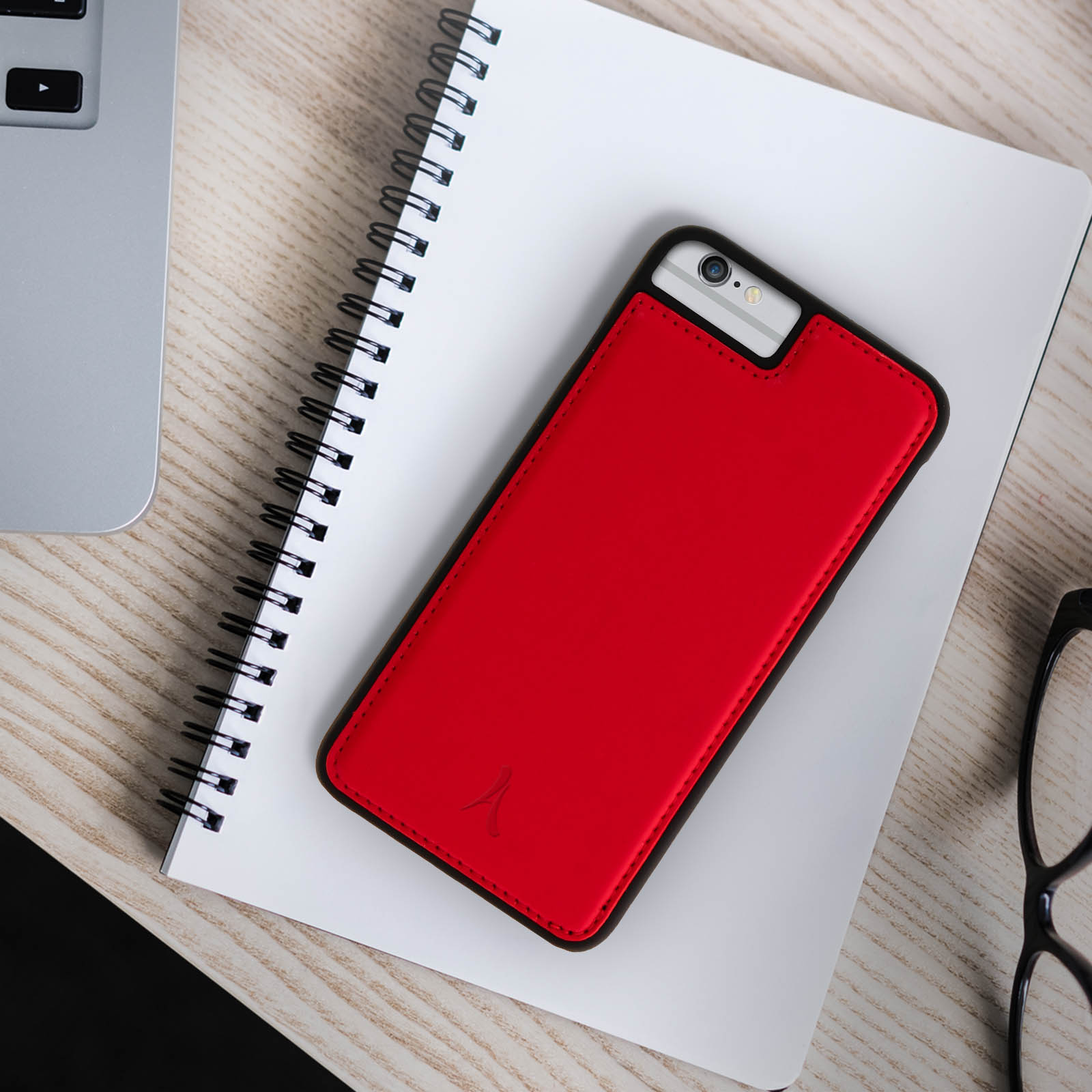 Custodia rossa per telefono su un taccuino. Un laptop e occhiali sono parzialmente visibili su un tavolo di legno.