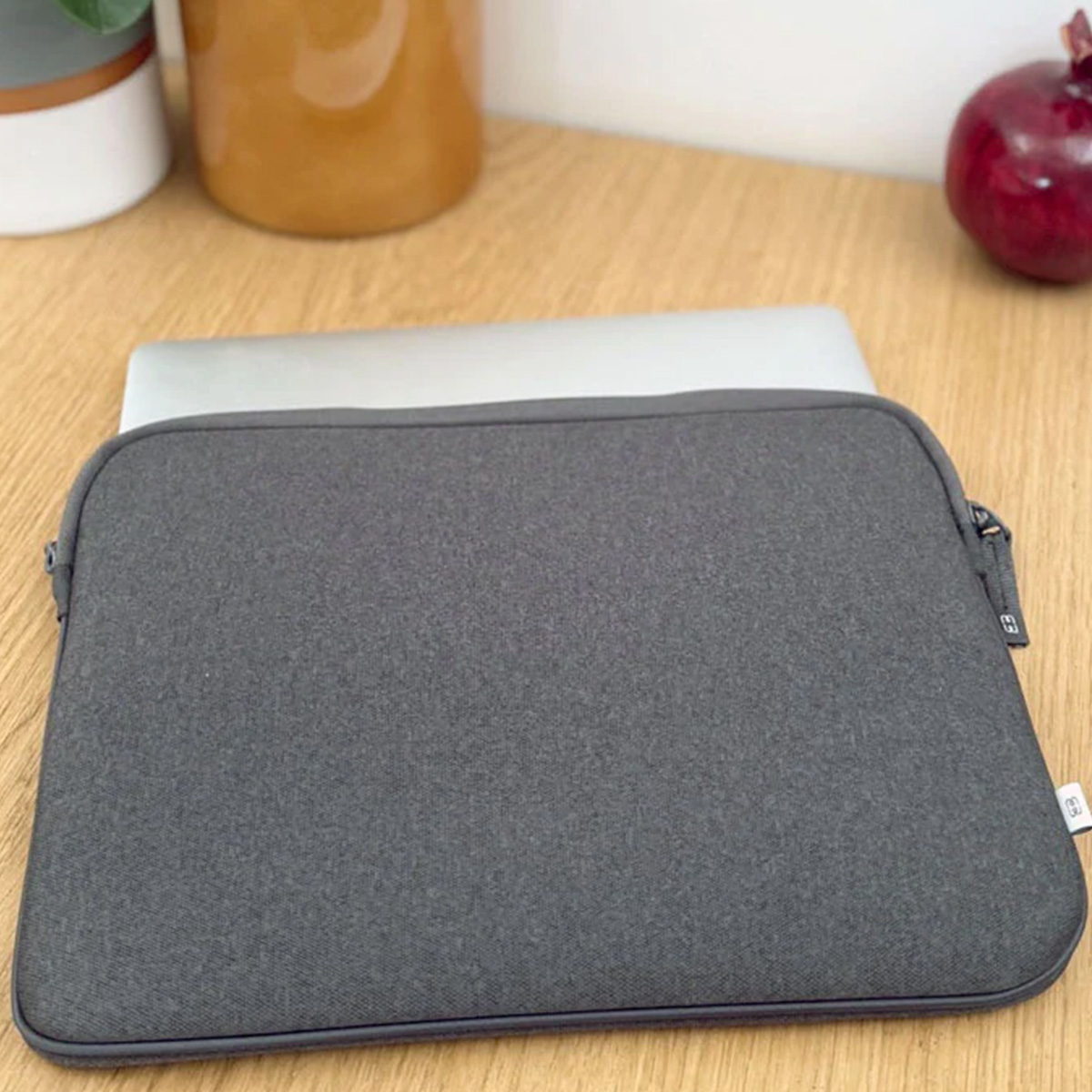 Una custodia grigia per laptop è su una superficie di legno. Un laptop è all'interno. Una mela rossa e delle piante sullo sfondo.