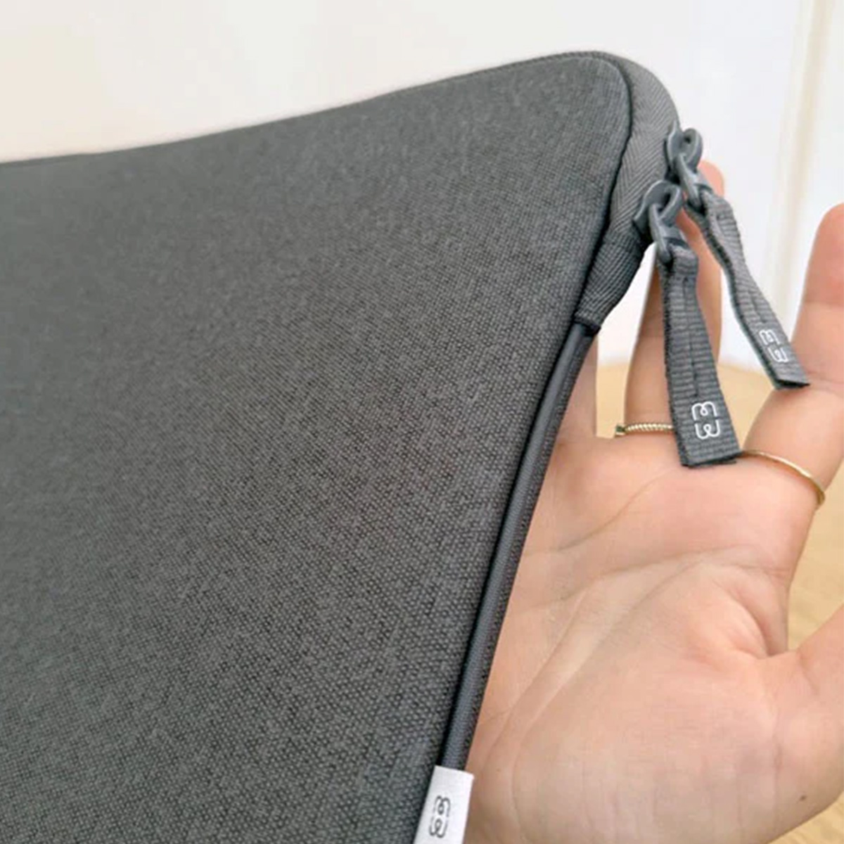 Una custodia grigia per laptop con cerniera è tenuta da una mano con anelli. Lo sfondo è chiaro.