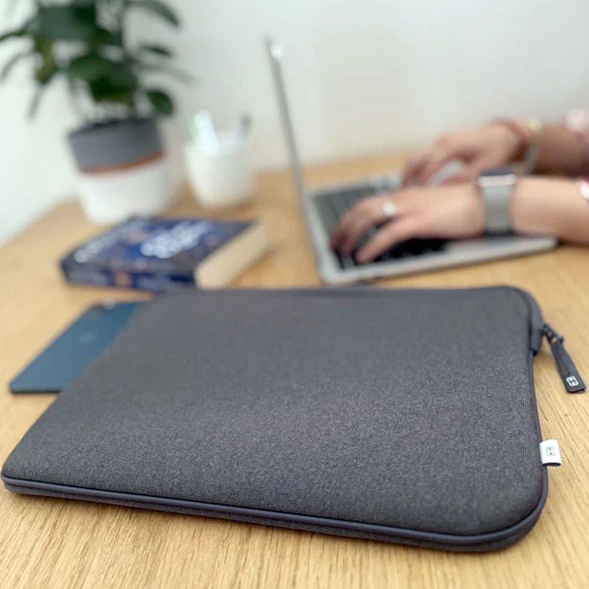 Custodia per laptop, laptop, libro, telefono e una persona che scrive su un laptop su una scrivania di legno. Una pianta sullo sfondo.