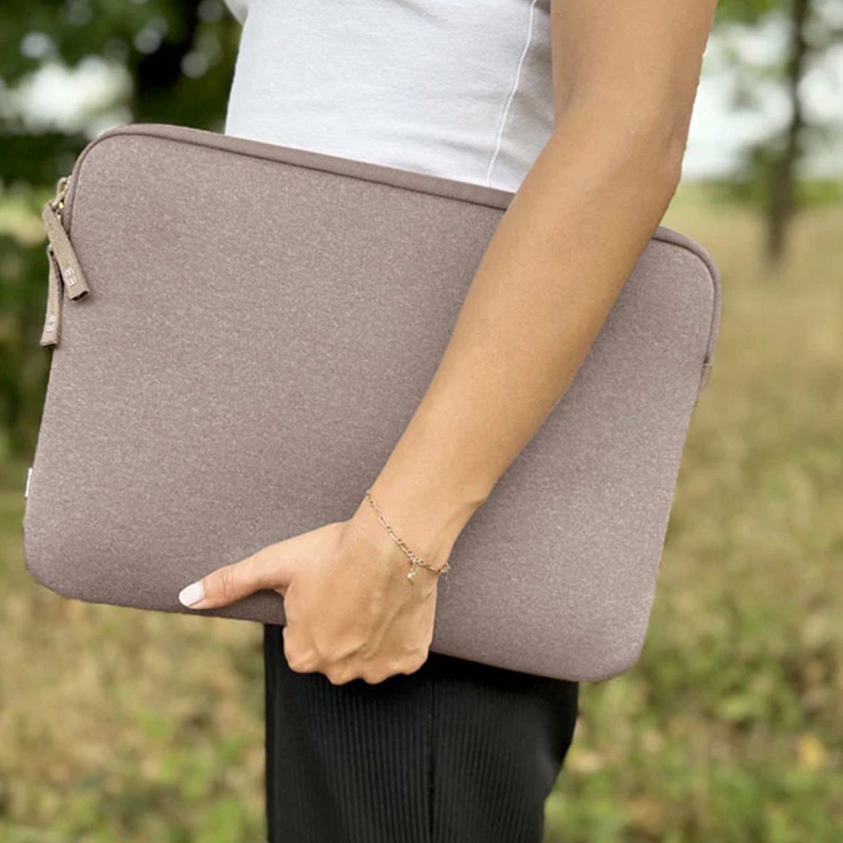 Una persona tiene in mano una custodia per laptop color talpa. Indossa una camicia bianca, pantaloni neri e un braccialetto. Fogliame verde sullo sfondo.