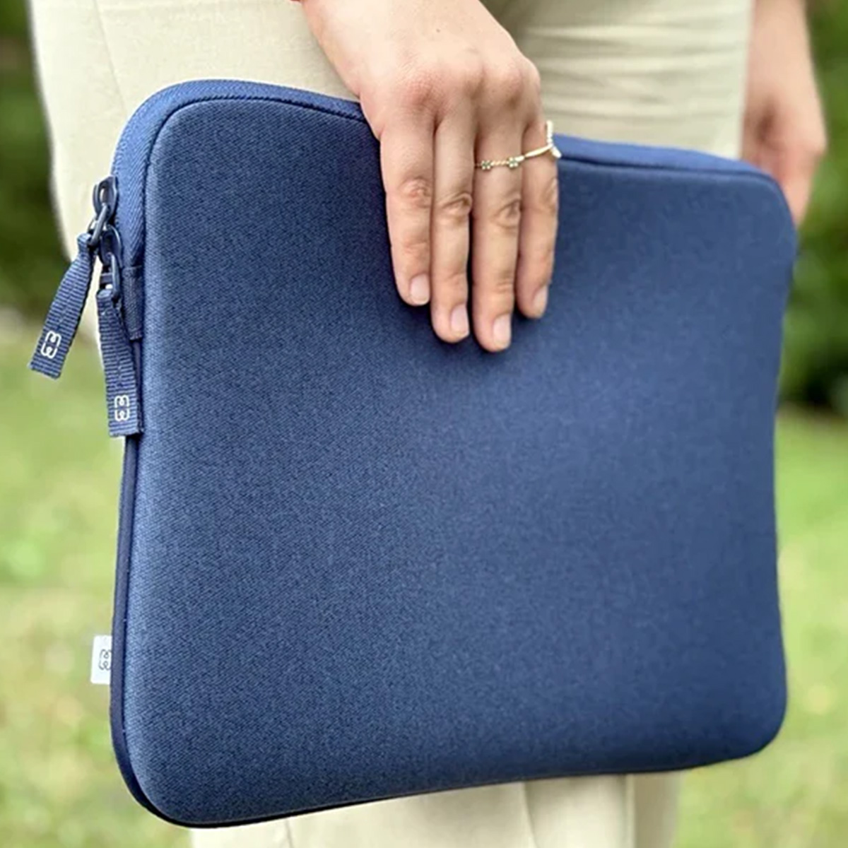 Una persona tiene in mano una custodia per laptop blu navy. La mano della persona con un anello è appoggiata sulla custodia. Sono visibili pantaloni beige.