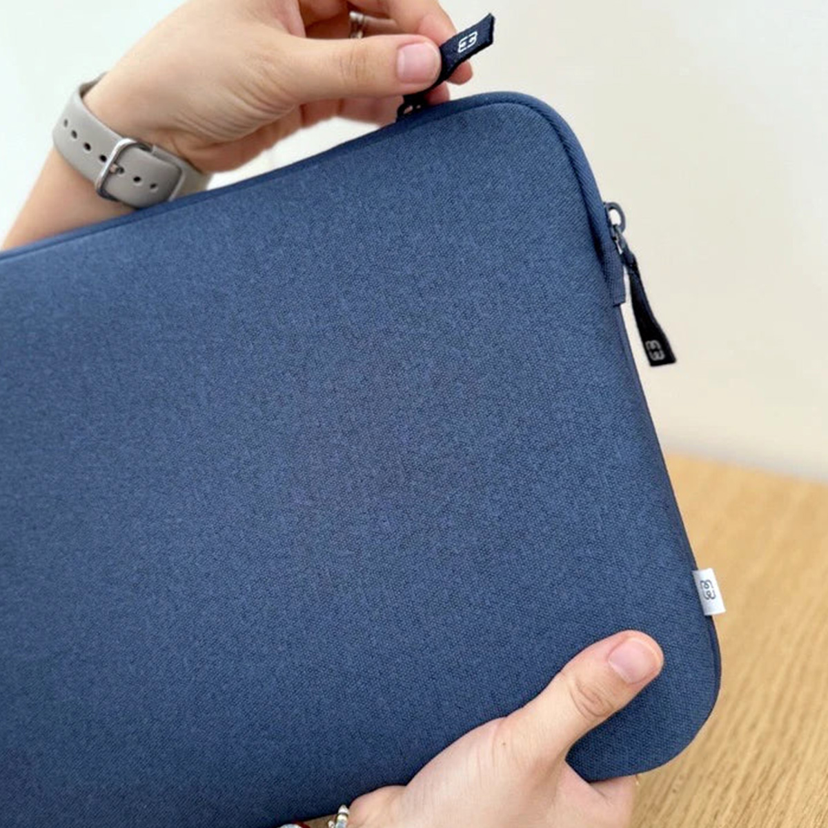 Mani che tengono una custodia per laptop blu navy con cerniera. Un orologio è al polso. Lo sfondo è un tavolo di legno e un muro bianco.