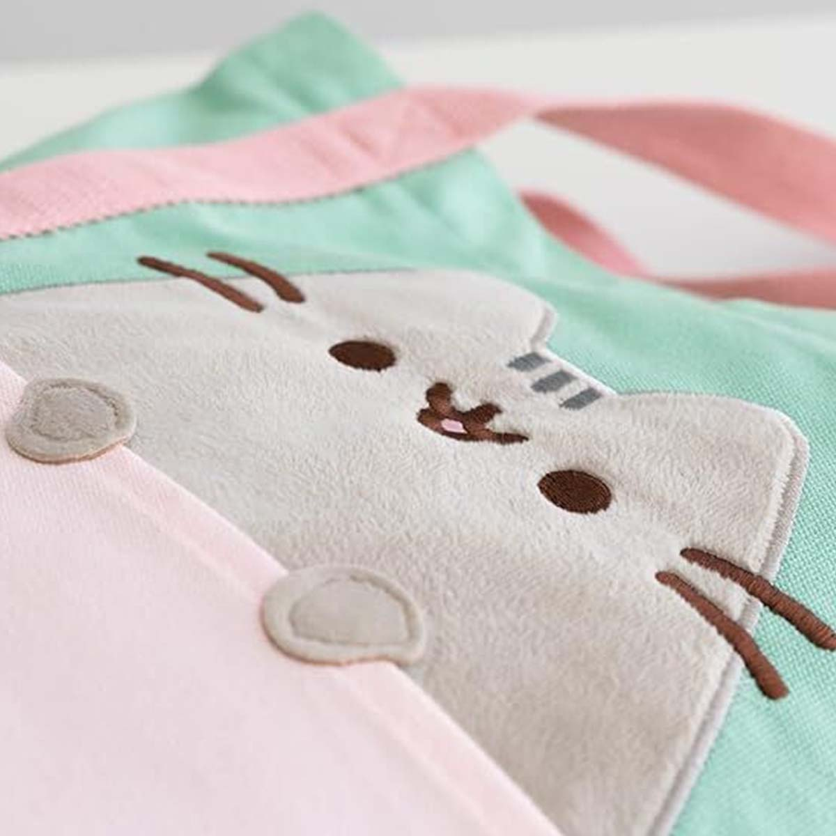 Un primo piano di una borsa a forma di gatto. La borsa è verde chiaro con bordo e manico rosa. Il volto del gatto è ricamato.