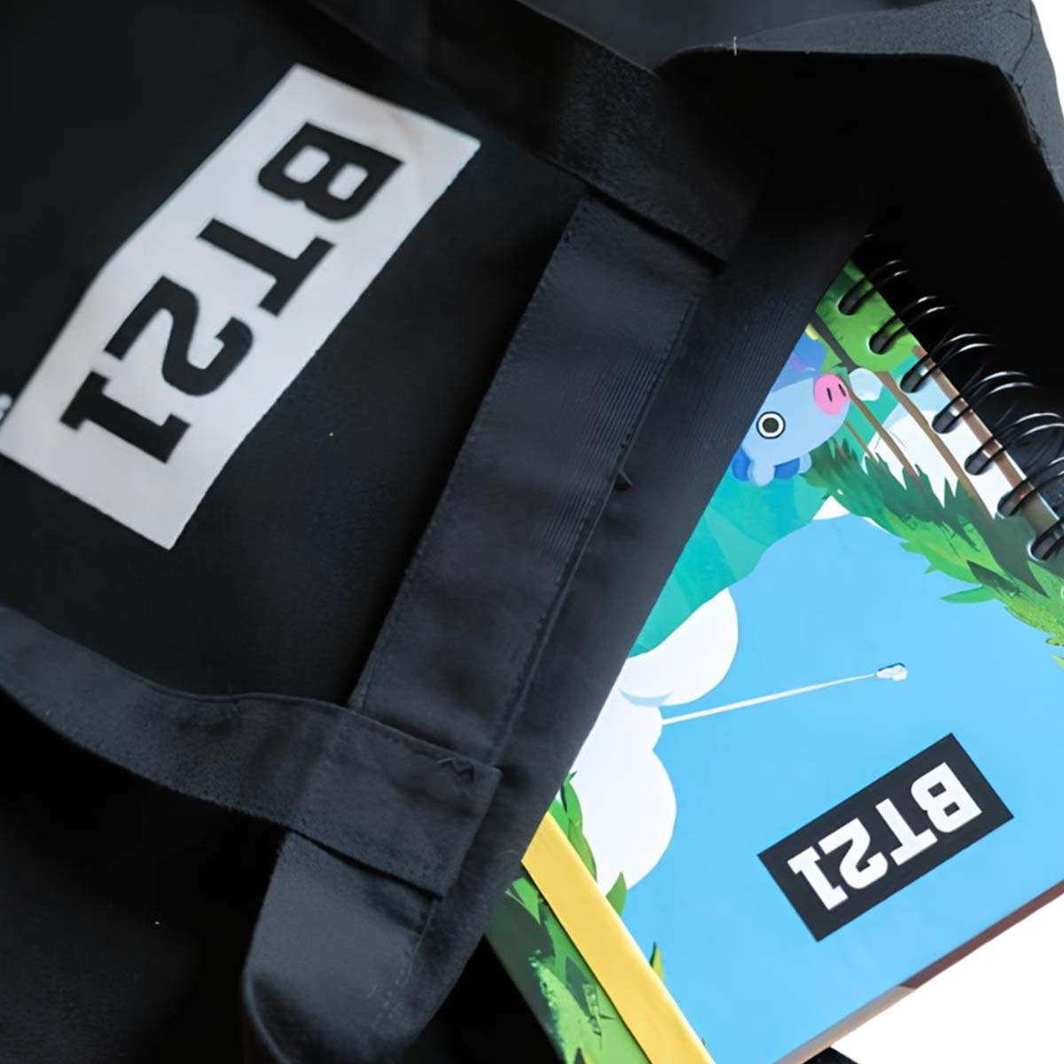 Borsa tote nera con logo BT21. All'interno, un quaderno a spirale con un personaggio dei cartoni animati e il logo BT21.