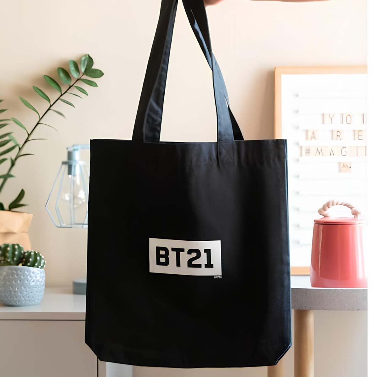 Una borsa tote nera con 'BT21' viene tenuta in mano. Sullo sfondo una pianta, una lampada, un cactus e un vaso rosa.