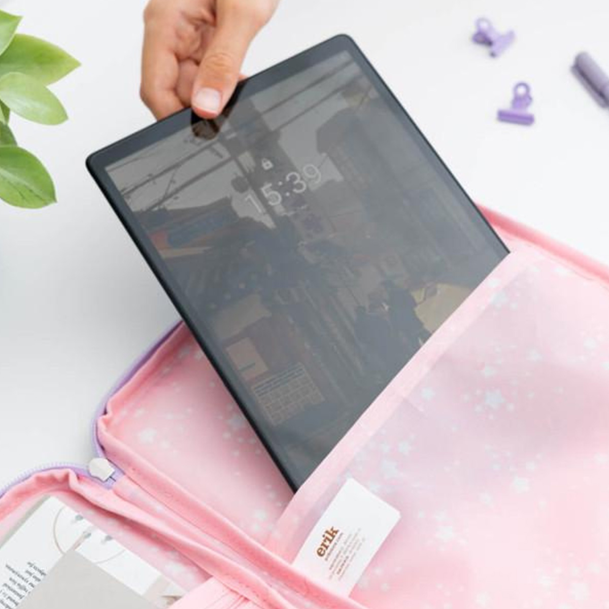 Mano che tiene tablet con display 15:39, in custodia rosa con stelle. Clip e penna viola visibili. Sfondo bianco.