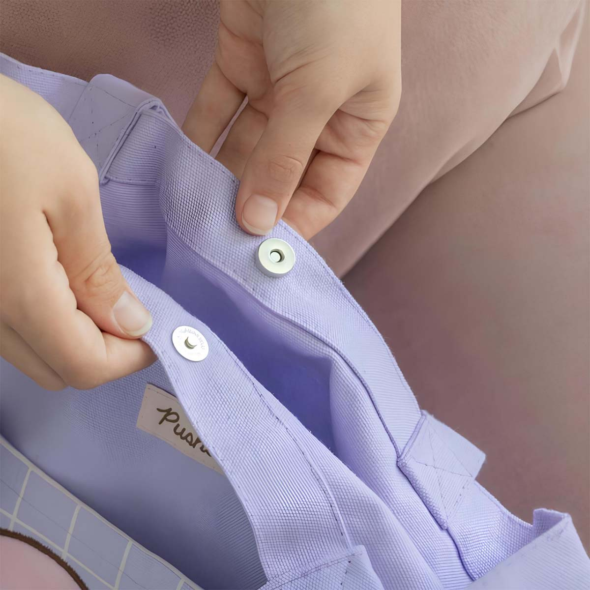 Mani che aprono una borsa tote viola. La borsa è tenuta da una persona che indossa abiti rosa. La borsa ha una chiusura magnetica.