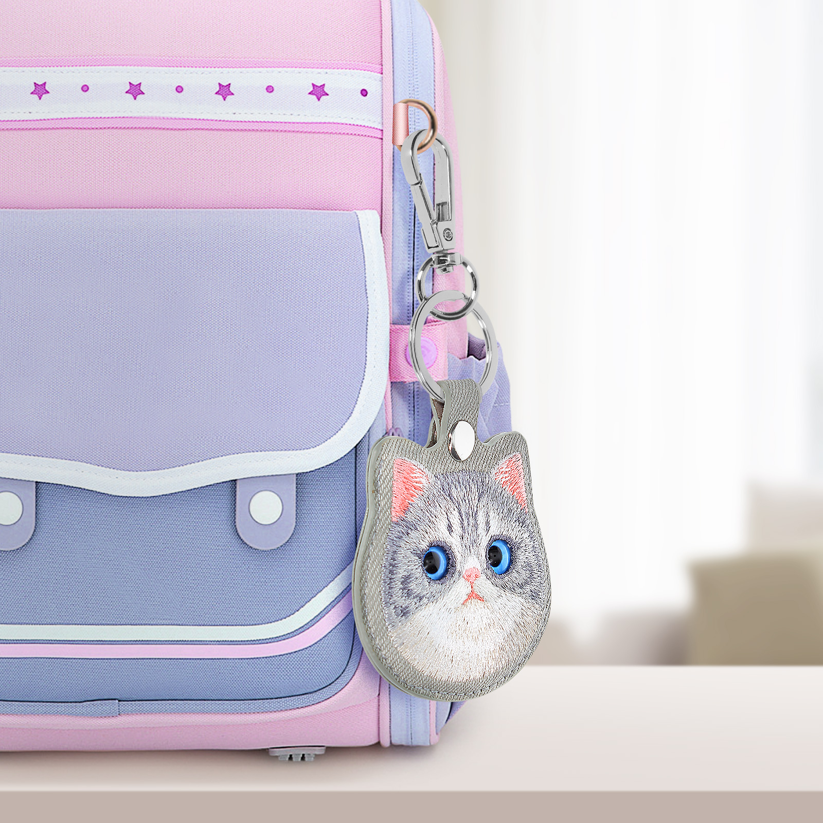 Un portachiavi a forma di gatto è appeso a uno zaino rosa, viola e blu. Il gatto ha occhi blu e pelliccia grigia.