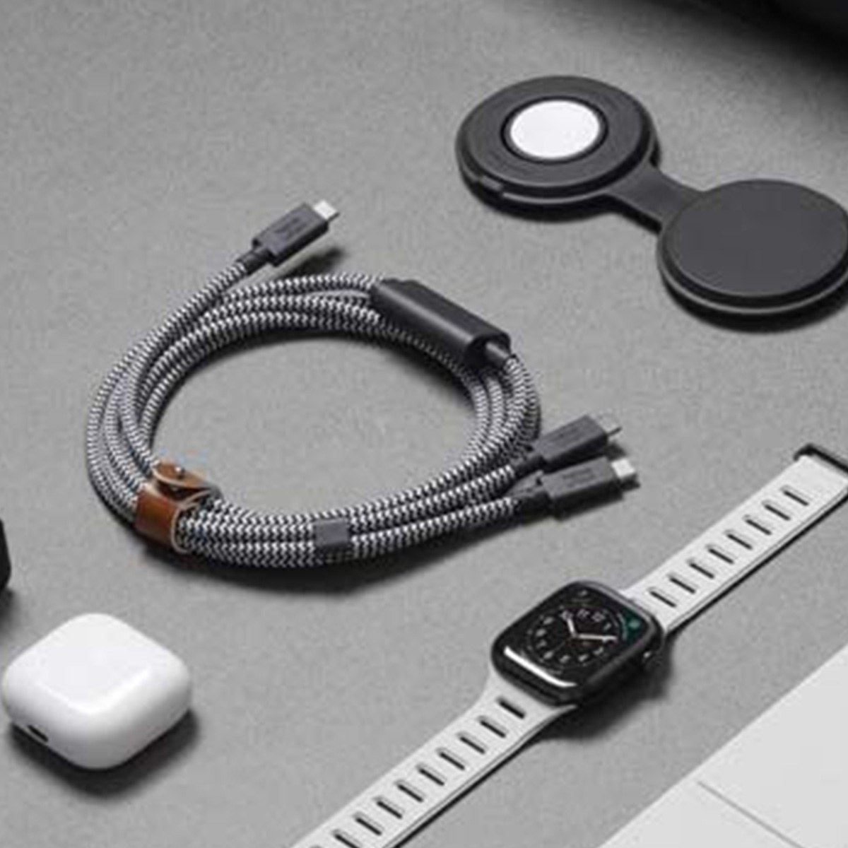 Draufsicht zeigt Tech-Zubehör: eine Uhr, Ladekabel, ein kabelloses Ladegerät und Ohrhörer. Alles auf einer grauen Oberfläche.