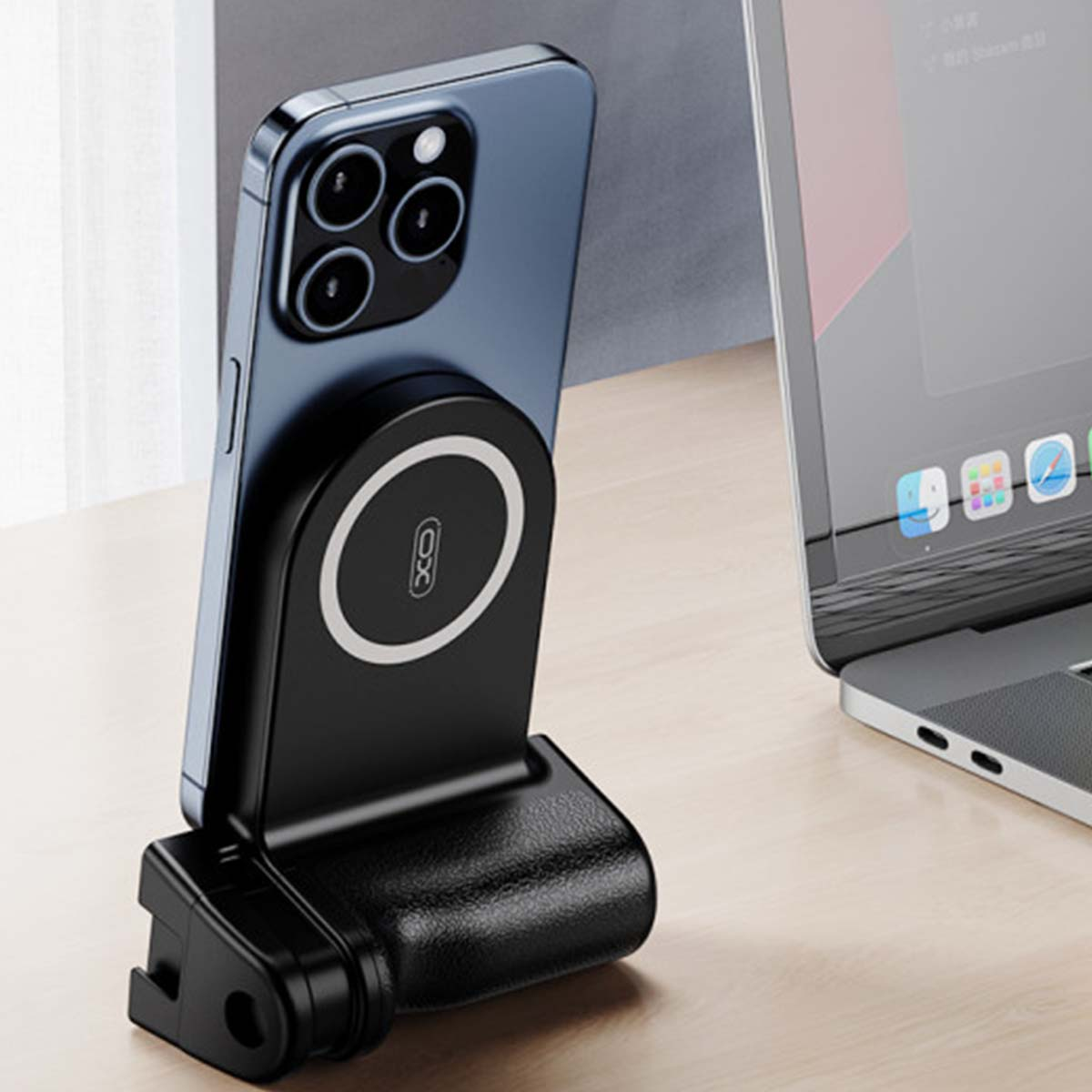 Un telefono è collegato a un caricatore wireless nero. Un laptop è sullo sfondo su una scrivania di legno. Una finestra è a sinistra.