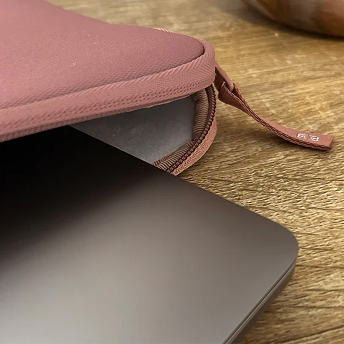 Un laptop parzialmente dentro una custodia rosa, su un tavolo di legno. La custodia ha una cerniera e un manico in tessuto. Il laptop è grigio scuro.