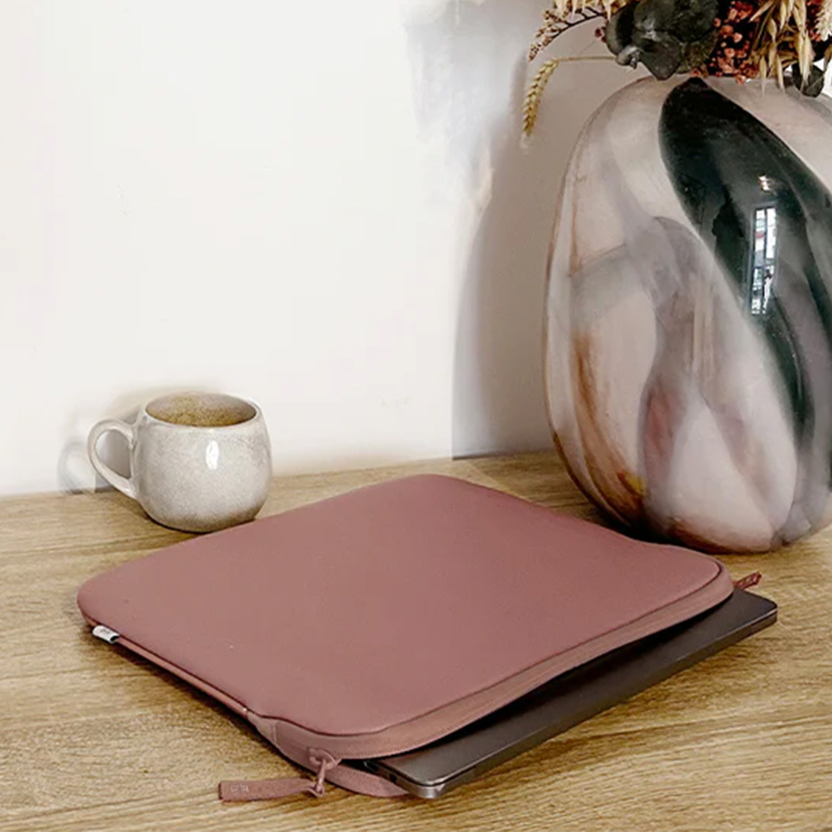 Un laptop in una custodia rosa è su un tavolo di legno. Una tazza e un vaso decorativo sono nelle vicinanze. Lo sfondo è un muro bianco.