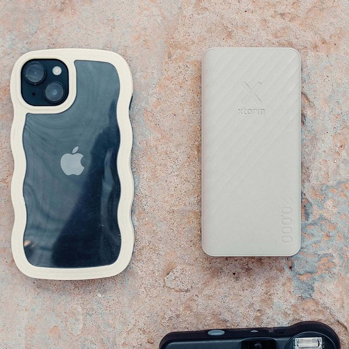 Un téléphone dans un étui, une batterie externe et un appareil photo sont sur une surface en pierre. Le téléphone a un logo Apple.